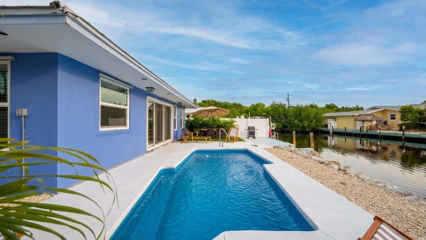 A swimming pool with a patio area, a house with blue exterior walls, and a calm water body in the background. - Dog-Friendly • Pool & Boat Dock • Sombrero Beach (Marathon)