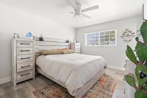 Een slaapkamer met een bed, een dressoir, een plafondventilator, een raam en een cactus.