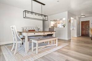 Een eethoek met een houten tafel, witte gestoffeerde stoelen en een bank. Daarboven hangt een moderne hanglamp.