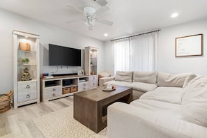 Een moderne woonkamer met een flatscreen televisie, een beige bank, een houten salontafel en een plafondventilator.