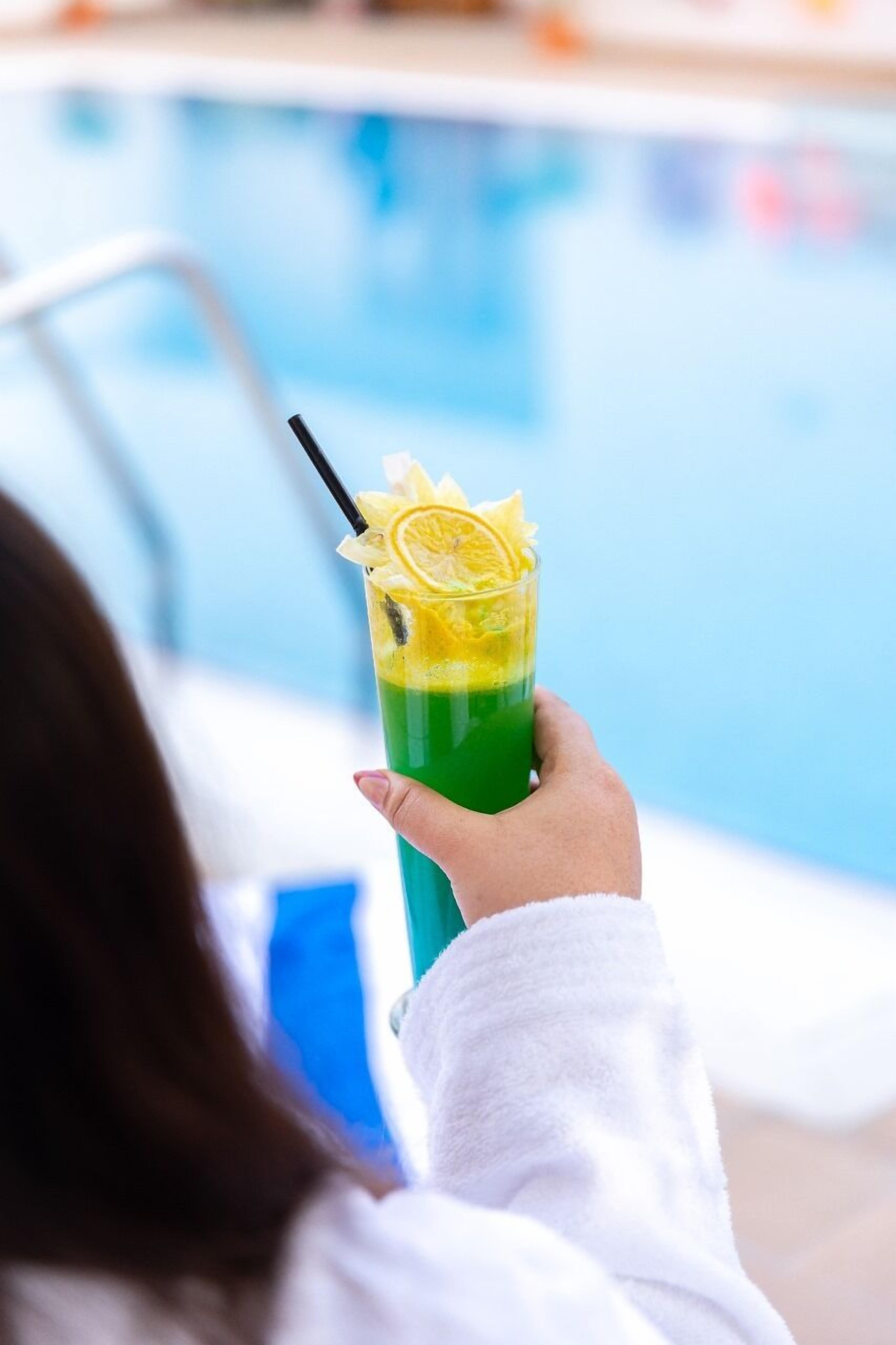 Una persona sostiene un brebaje verde con pajita y decoración de limón junto a una piscina.