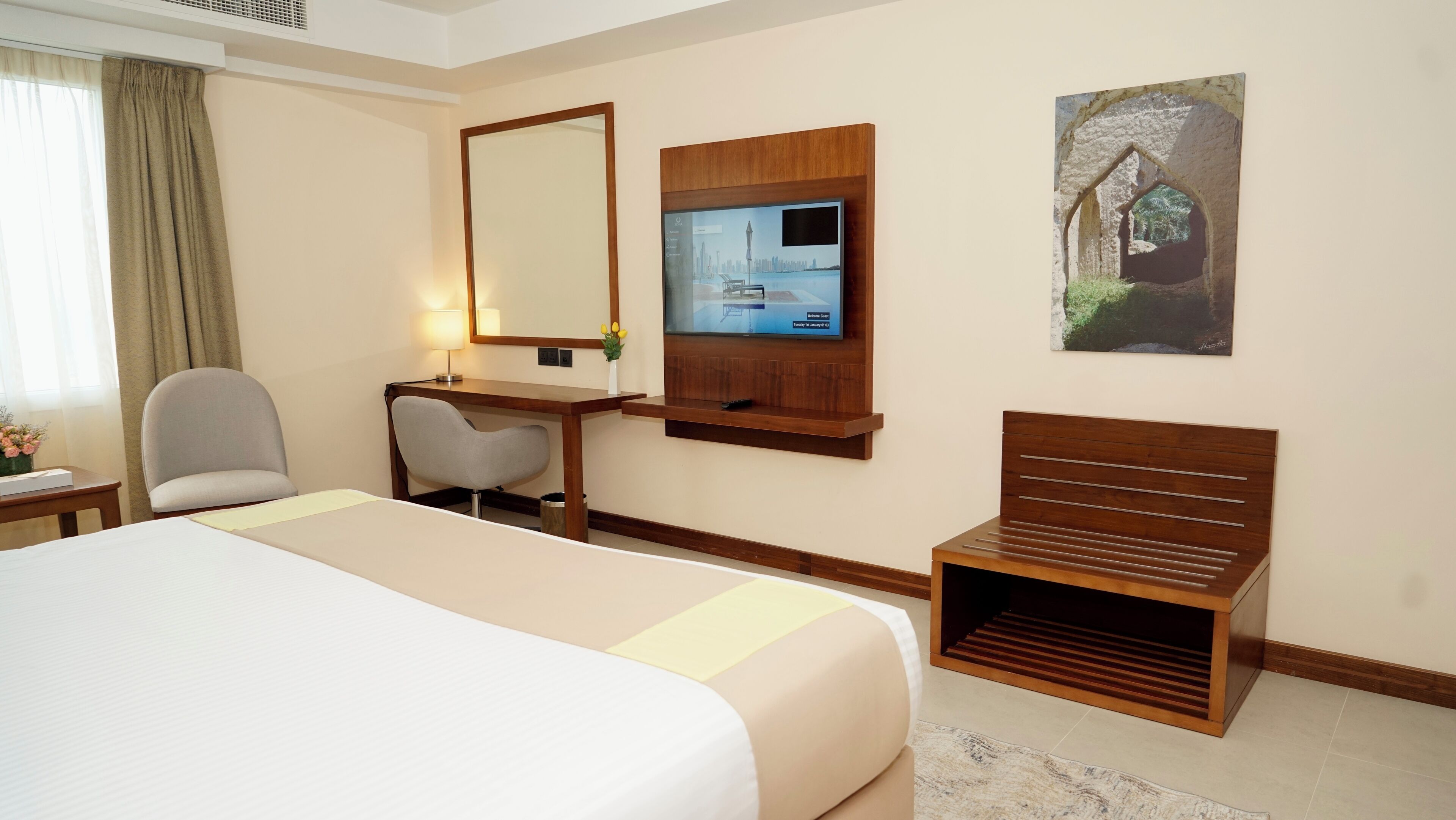 A hotel room with a bed, a desk, a chair, a TV mounted on the wall, and a small bench.