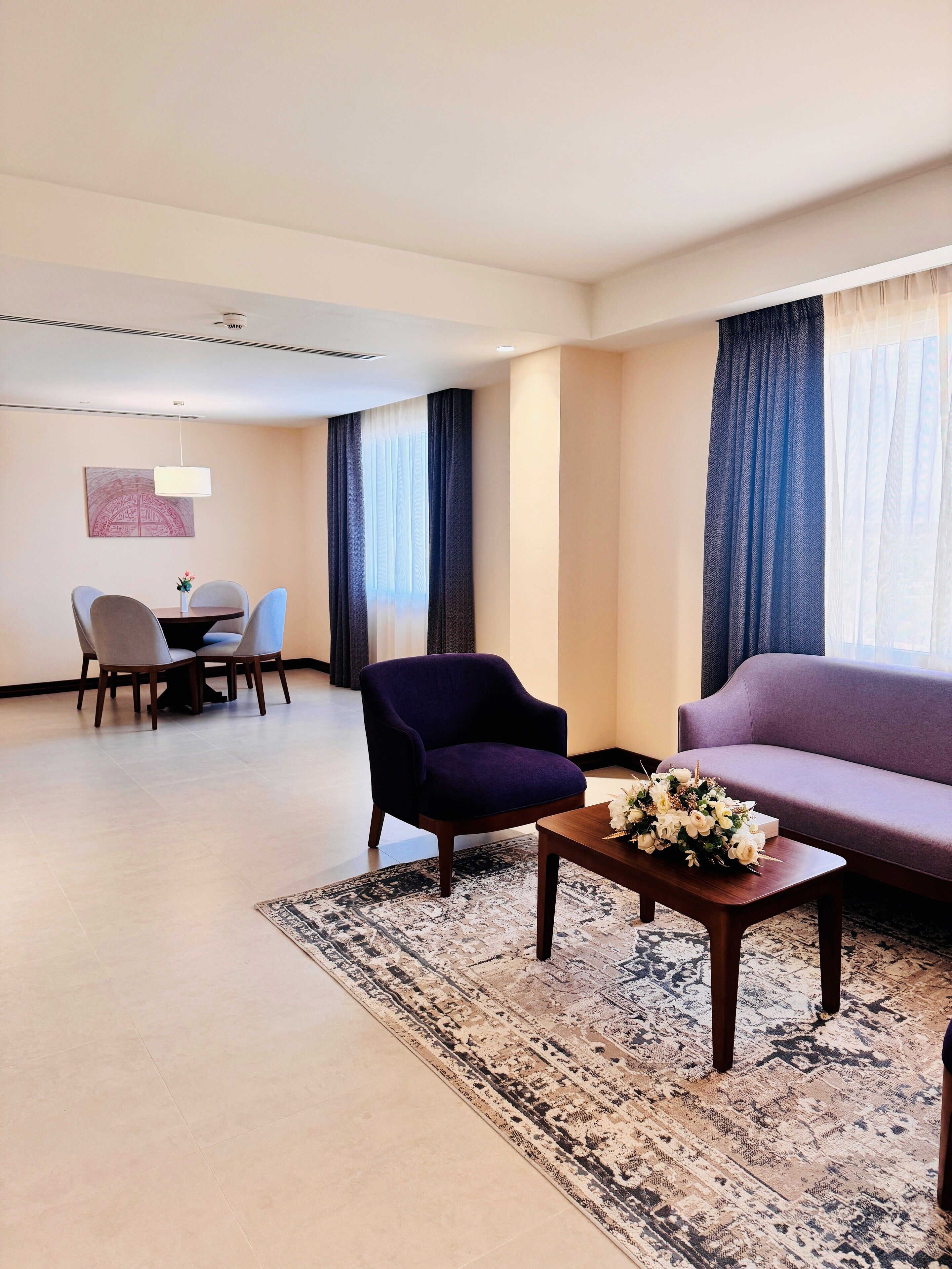 Habitación de hotel con un sillón púrpura, un sofá, una mesa de comedor con sillas y una mesa de centro con flores.