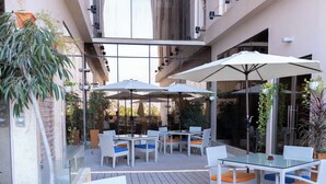 Een terras buiten met witte rieten meubels, glazen tafels en oranje kussens.