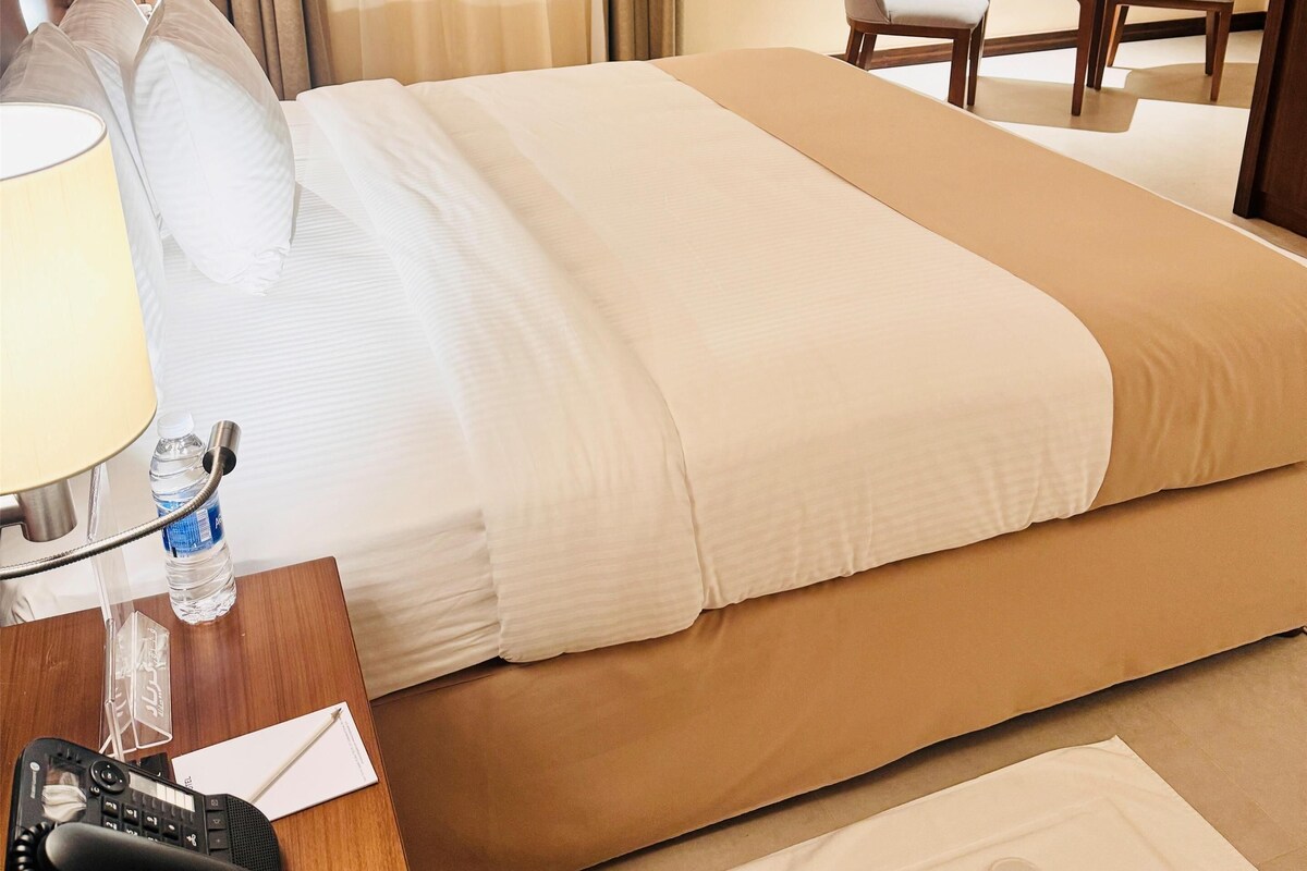 Una habitación de hotel con una cama, un escritorio con teléfono, una silla, una mesa y un cuadro en la pared.