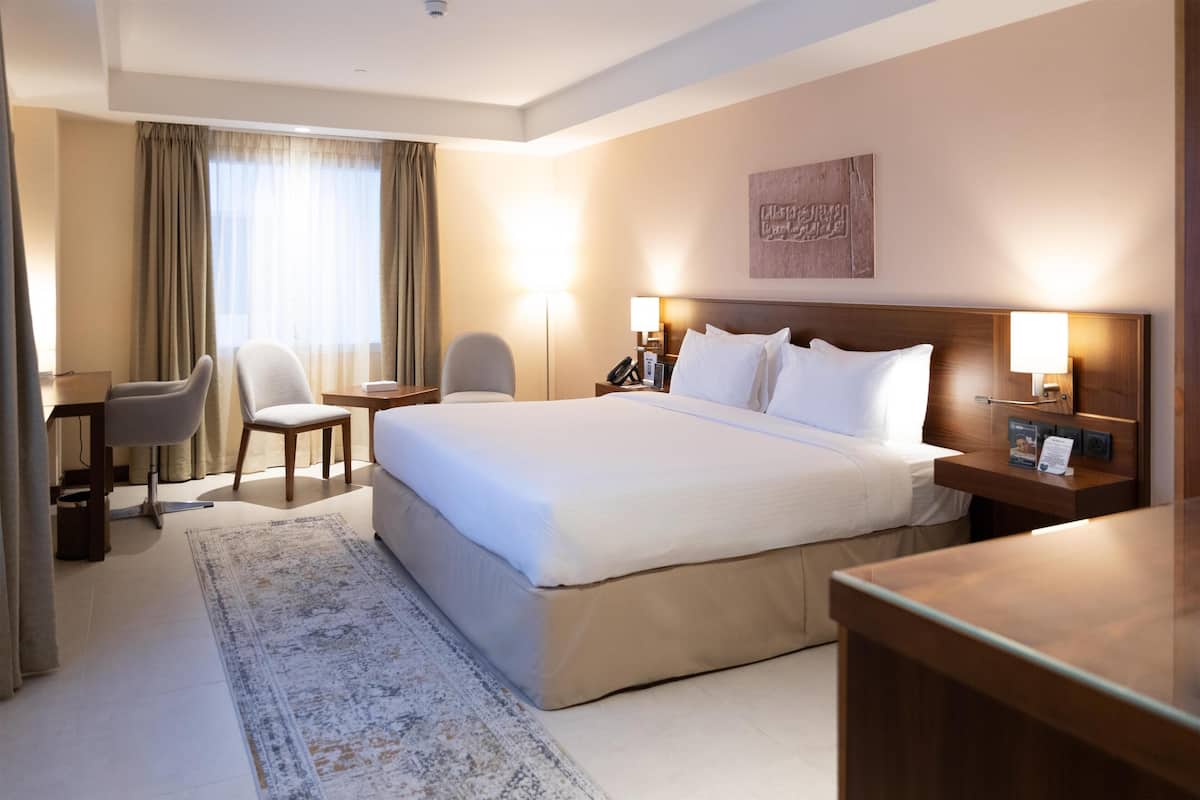 Hotelkamer met een groot bed, twee nachtkastjes, een bureau, een stoel en een groot raam met gordijnen.