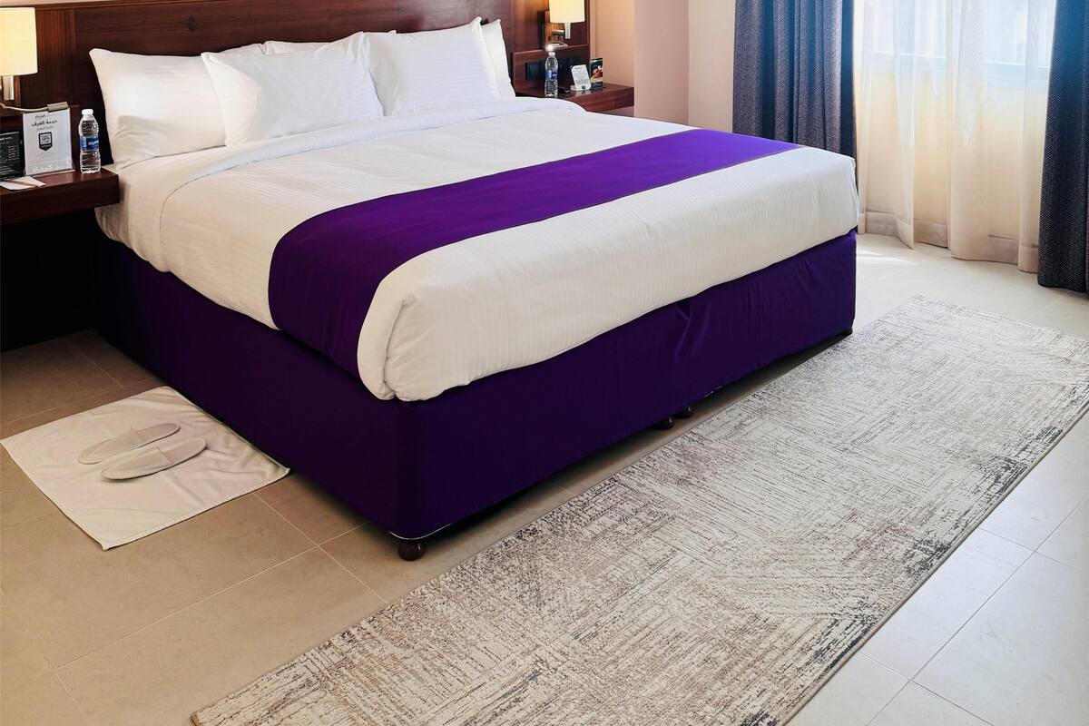 Een hotelkamer met een groot bed, een hoofdeinde met een schilderij, een nachtkastje met een lamp en een raam met gordijnen.