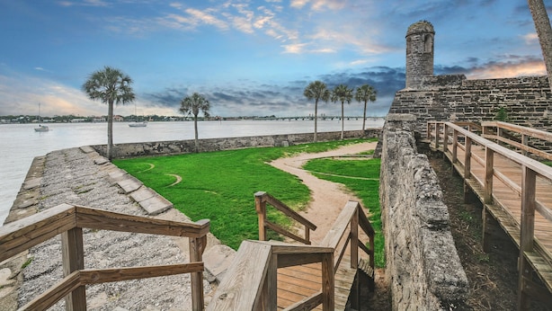 Un paseo marítimo con muro de piedra, barandas de madera y una zona verde que conduce al agua.