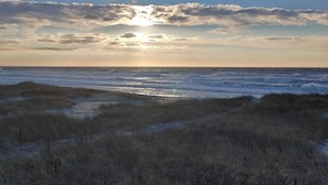 A coastal landscape with a path leading through low-lying vegetation, a clear sky, and the sun setting or rising over the horizon. - THE SANDBAGGER @ OCEAN ISLE (Ocean Isle Beach)