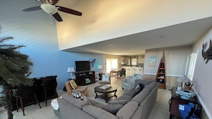 A spacious living room with a large sectional sofa, a ceiling fan, and a dining area with a kitchen in the background. - THE SANDBAGGER @ OCEAN ISLE (Ocean Isle Beach)