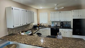 A modern kitchen with white cabinets, granite countertops, and a central island with stools. - THE SANDBAGGER @ OCEAN ISLE (Ocean Isle Beach)