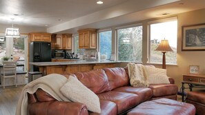 A living room with a brown leather couch, a dining area with a table and chairs, a kitchen with wooden cabinets, and a large window with a view of trees. - Stunning Panoramic Views of Mtns & Lake (Eden)