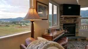 Un salon chaleureux avec une cheminée, un téléviseur fixé au-dessus et une vue sur l’extérieur.