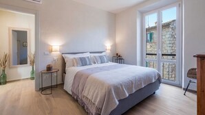 Un dormitorio con cama, mesitas de noche, un espejo y vistas a un edificio de piedra a través de una ventana.
