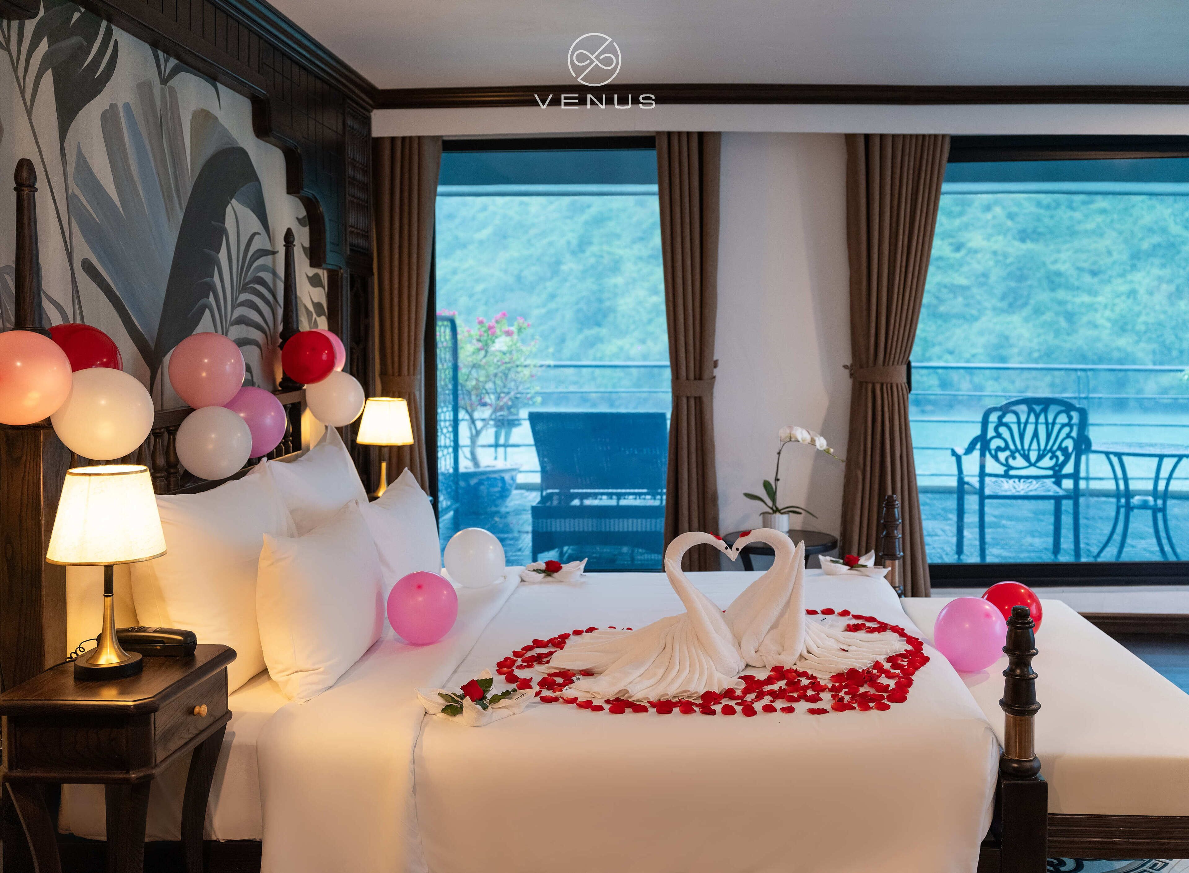 A hotel room with a bed, a nightstand, a lamp, a bouquet of roses, and a view of the outdoors.