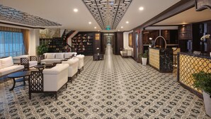 Spaziosa hall con pavimento a motivi, area salotto con divani e tavolini, reception e scala che conduce ai piani superiori.