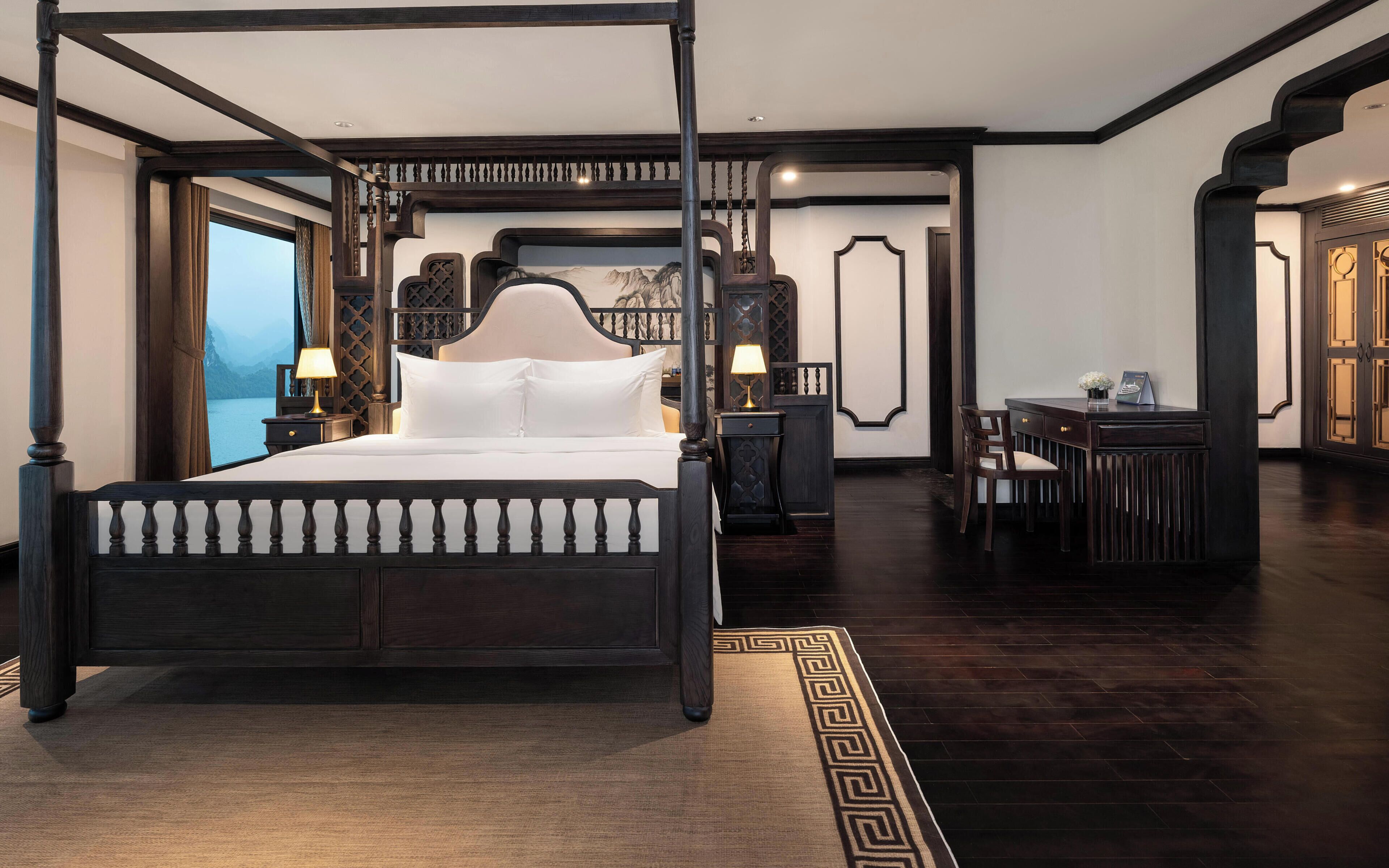 A four-poster bed with a headboard, bedside tables with lamps, a desk with a chair, and a view of a body of water through a window.