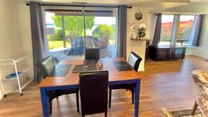 Un comedor con una mesa de madera, sillas y una computadora portátil. Se ve un jardín a través de ventanas amplias.