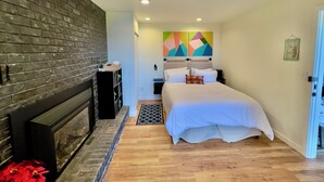 Un dormitorio con pared de ladrillo, cama con ropa de cama blanca, mesita de noche, televisor montado en la pared y un cuadro abstracto colorido sobre la cama.