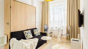 Une chambre d’hôtel moderne avec un canapé noir, une armoire en bois, une table à manger avec des chaises, un téléviseur fixé au mur et une grande fenêtre avec des rideaux.