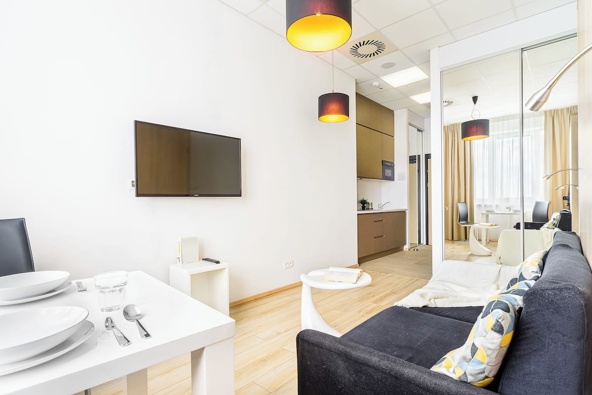 A modern living room with a sofa, a dining table with plates and cutlery, a TV, and a kitchen in the background.