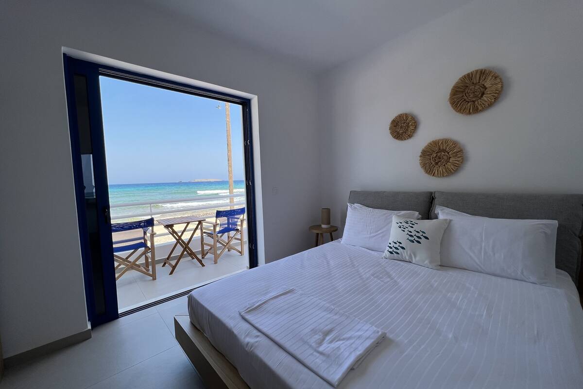 A bedroom with a bed, a view of the beach, and a balcony with chairs.