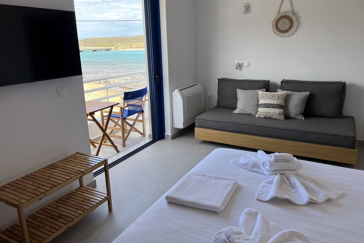 A modern hotel room with a bed, a sofa, a TV, a view of the beach, and a wooden chair.