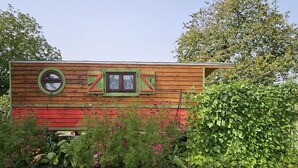 A wooden house with a red and green exterior, a window with a green frame, and a garden with various plants and flowers. - ROULOTTE ANAÏS A LA CHAUM'YER (GRANDCOURT)