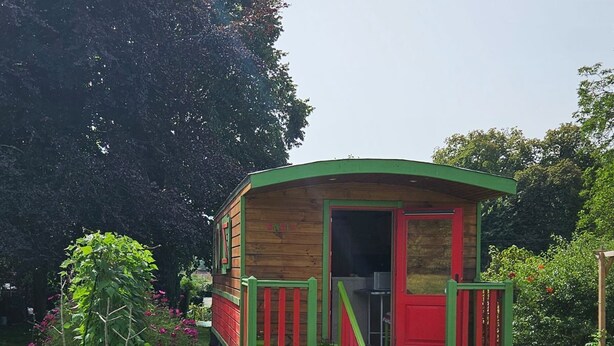 A small wooden cabin with a red door and green accents, situated in a grassy area with a potted plant nearby. - ROULOTTE ANAÏS A LA CHAUM'YER (GRANDCOURT)