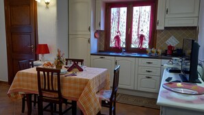 A kitchen with a dining table set for two, a window with decorative curtains, and a countertop with various kitchen items. - Apartment I Gelsomini # (Tanaunella)