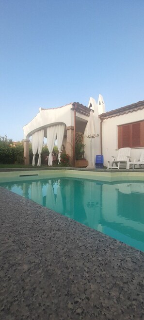 A swimming pool with a pergola and umbrellas, a covered seating area, and a building with a tiled roof. - Apartment I Gelsomini # (Tanaunella)