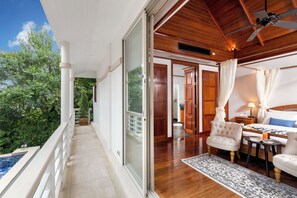 Ein geräumiger Balkon mit Blick auf üppiges Grün, Holzboden und ein modernes Schlafzimmer mit Bett, Nachttisch und kleinem Tisch mit Lampe.
