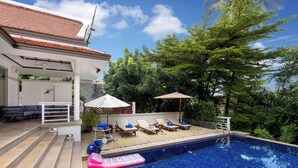 Una piscina con sillas de descanso y sombrillas, rodeada de árboles y un edificio con tejado de azulejos.