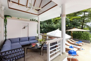 A covered outdoor seating area with a blue sofa, a table with a vase and a book, and sun loungers with towels.