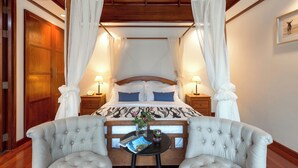 Una cama con dosel y cortinas blancas, cabecera de madera y mesita de noche con una revista y un florero.