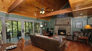 A cozy living room with a fireplace, wooden ceiling beams, and a large window overlooking a deck. - A River Runs Through It: Right on the River (Sevierville)