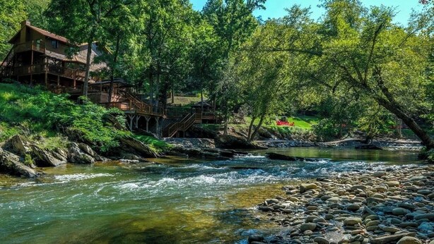 A riverside lodge with a wooden deck and a bridge, surrounded by lush greenery. - A River Runs Through It: Right on the River (Sevierville)