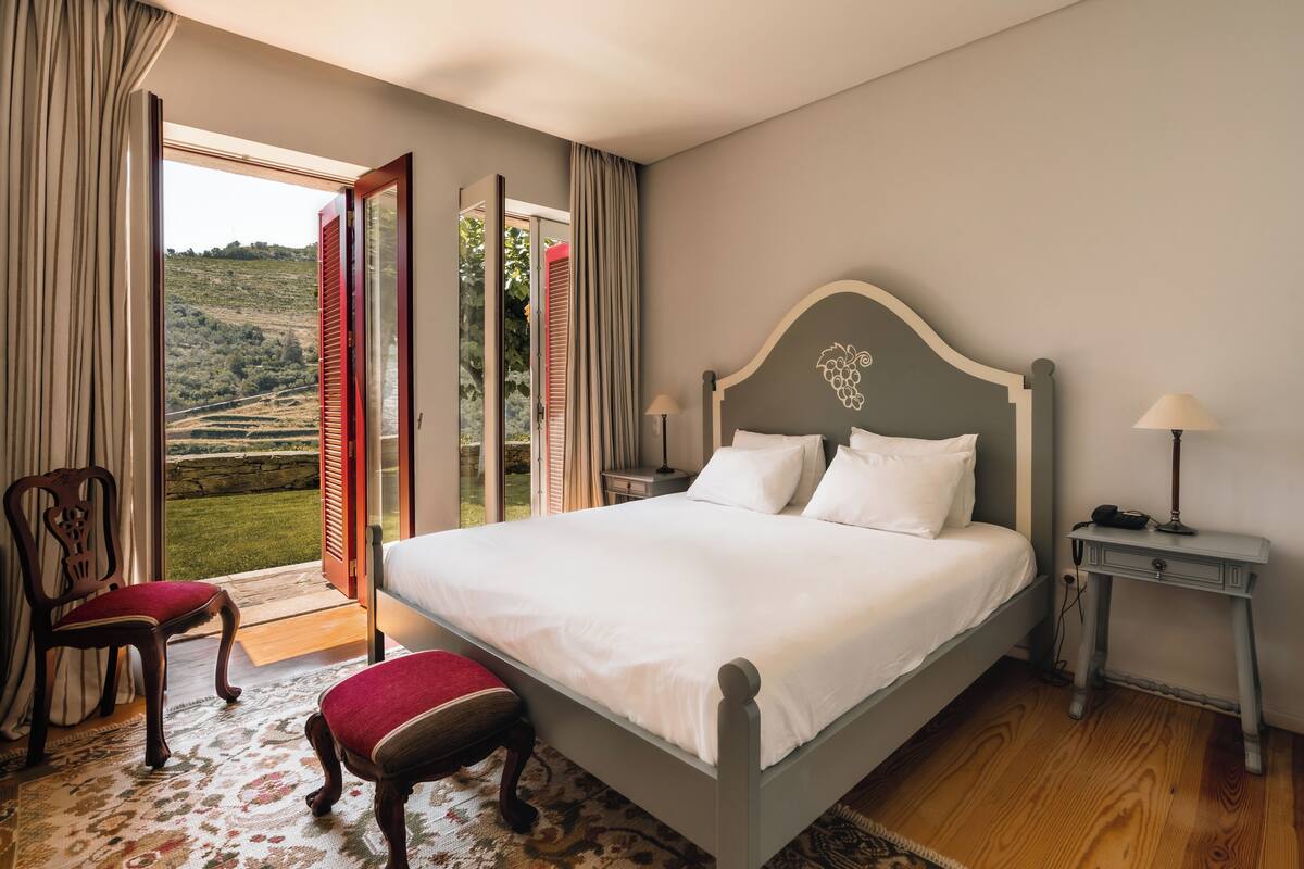 A bedroom with a large bed, a chair, a nightstand, and a view of a garden through an open door.