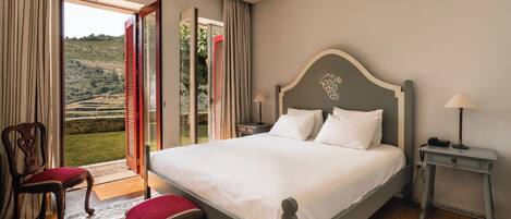 A bedroom with a large bed, a chair, a nightstand, and a view of a garden through an open door.