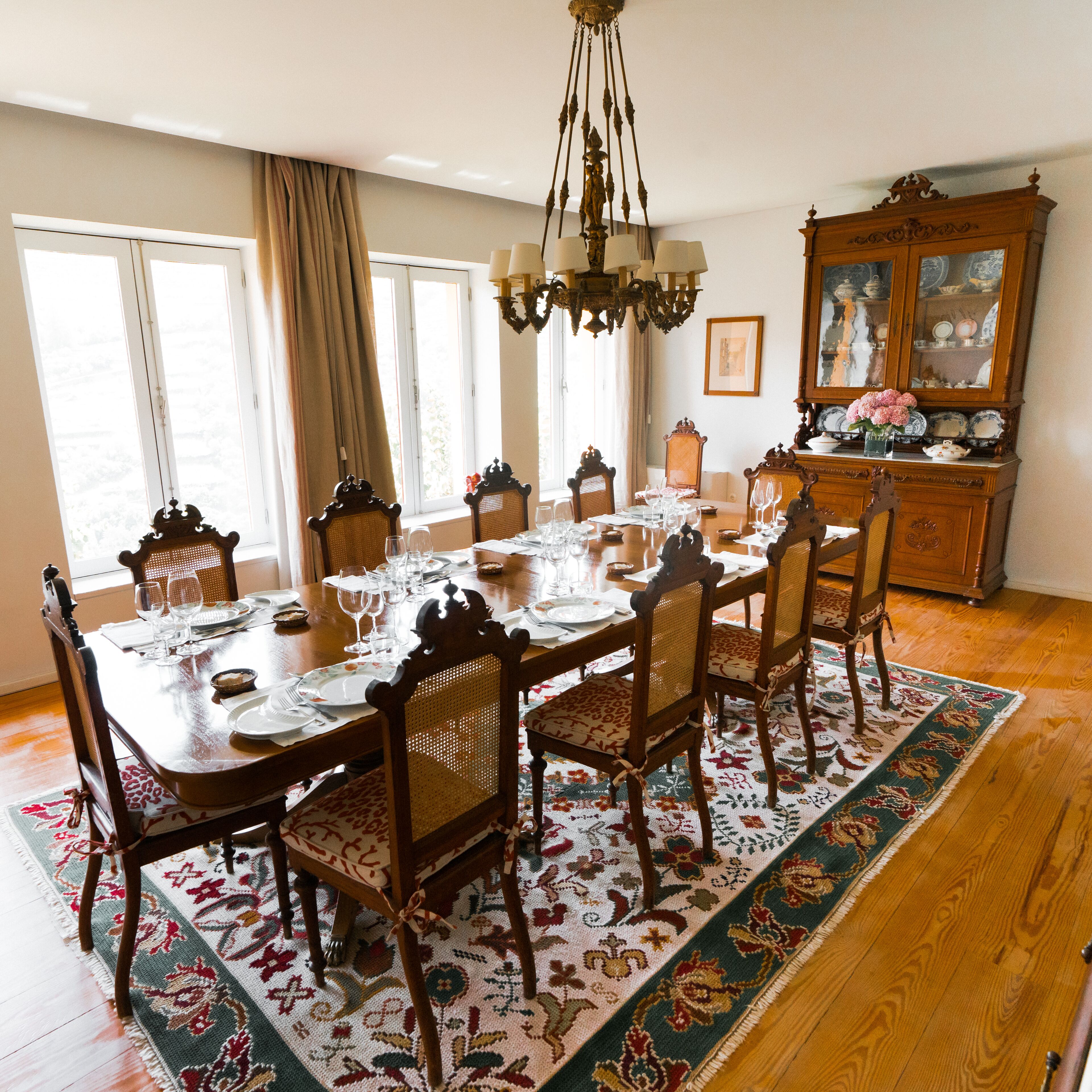 A dining room with a large wooden table set for six, surrounded by six chairs with woven backs. A chandelier hangs from the ceiling, and a wooden cabinet with glass doors is visible in the background.
