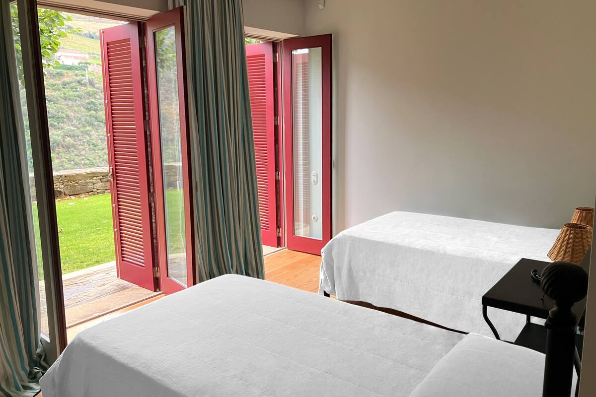 A bedroom with two beds, a wooden floor, and a view of a garden through open red shutters.