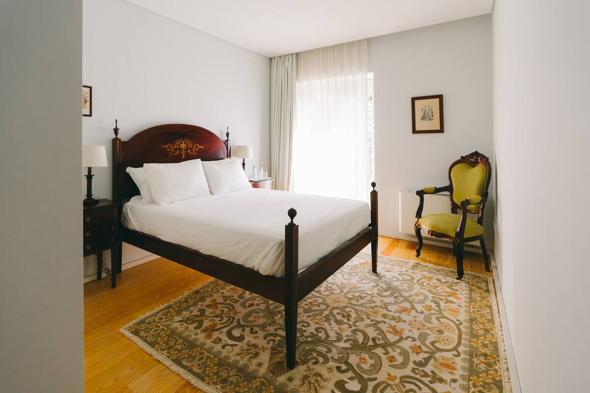 A neatly made bed with a wooden headboard, a green chair, a nightstand with a lamp, and a patterned rug on a wooden floor.