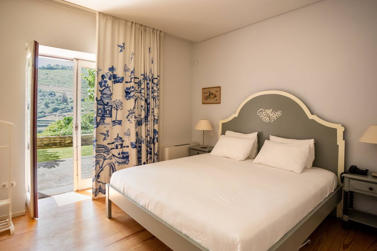 A bedroom with a bed, bedside tables, a nightstand, a window with curtains, and a view of greenery outside.