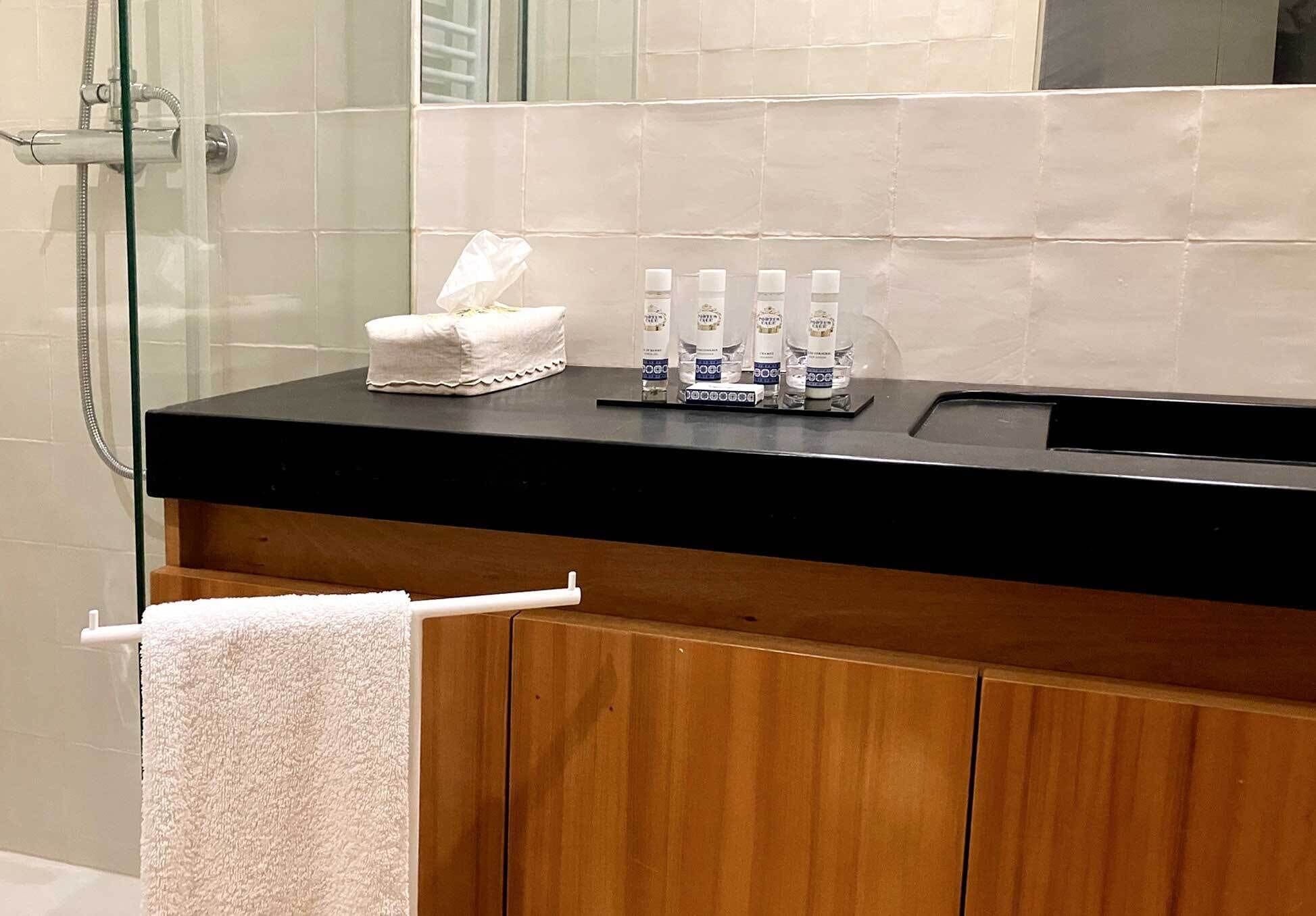 Un baño con encimera negra, un gabinete de madera debajo, cerramiento de ducha de vidrio y una toalla blanca colgada en un perchero.