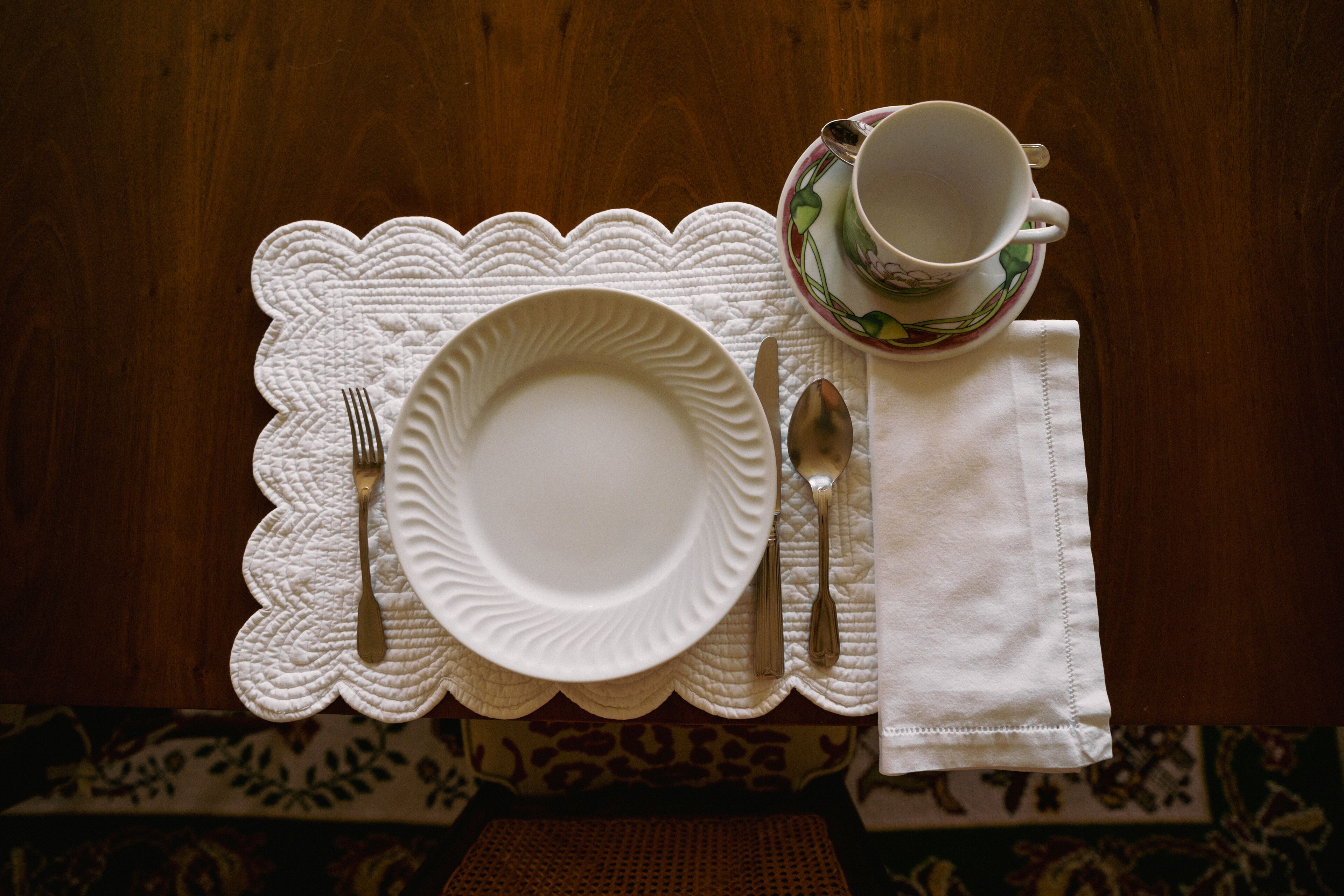 A neatly set table with a white plate, fork, knife, and spoon, accompanied by a cup and saucer.