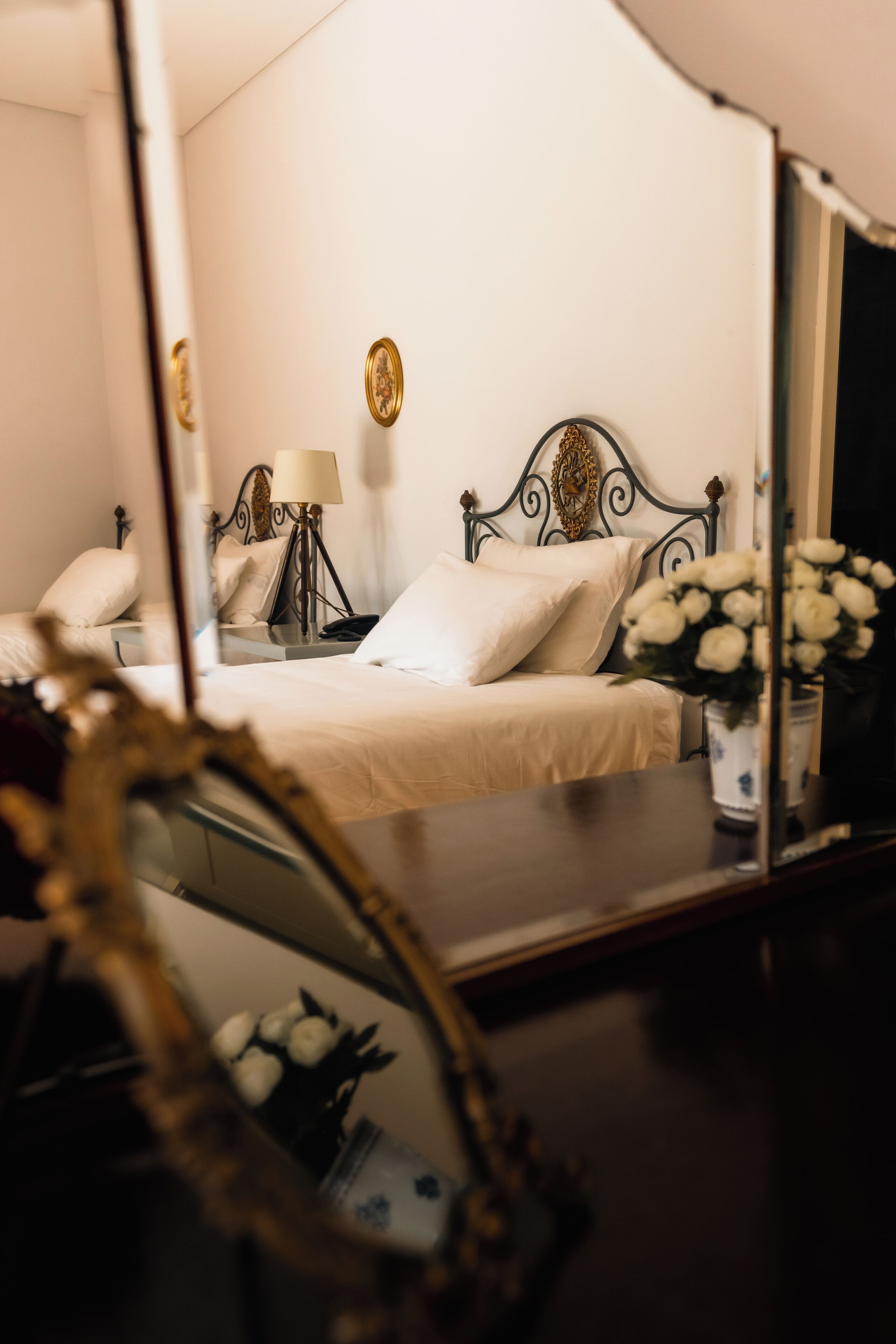 Quarto com cama, mesa de cabeceira, abajur e espelho refletindo o ambiente.