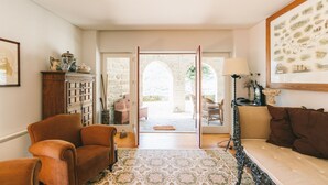 Ein gemütliches Zimmer mit einer Couch, Sesseln, einem gemusterten Teppich und Blick durch eine offene Tür auf einen Außenbereich mit einer Steinmauer und viel Grün.