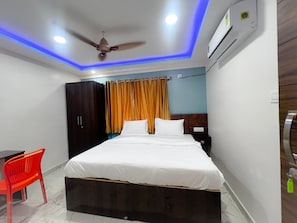Un dormitorio con cama, ventilador de techo, equipo de aire acondicionado, mesita de noche, una silla roja y una cortina.