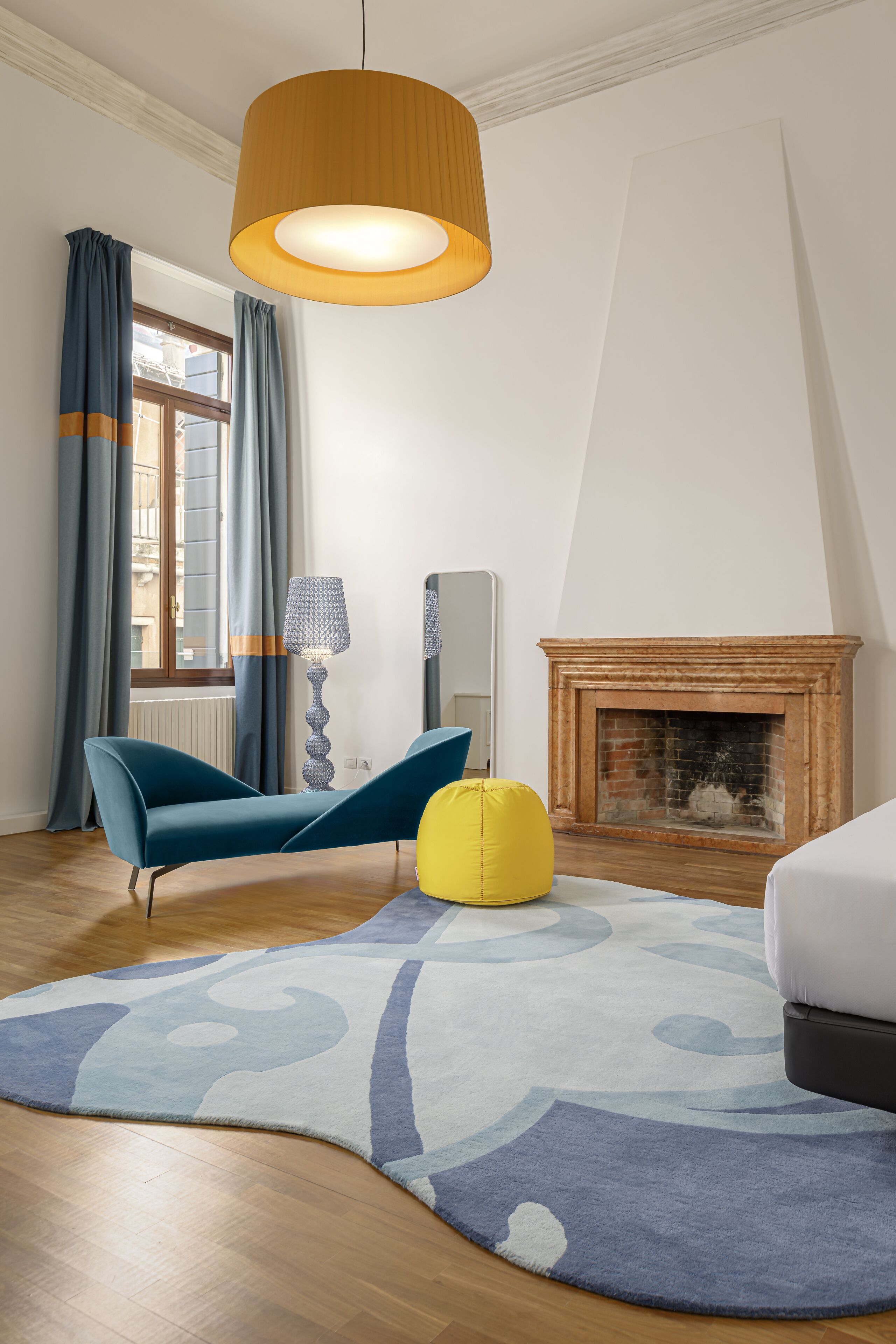 A modern room with a wooden floor, a blue sofa, a yellow bean bag, a patterned rug, a fireplace, and a hanging lamp.