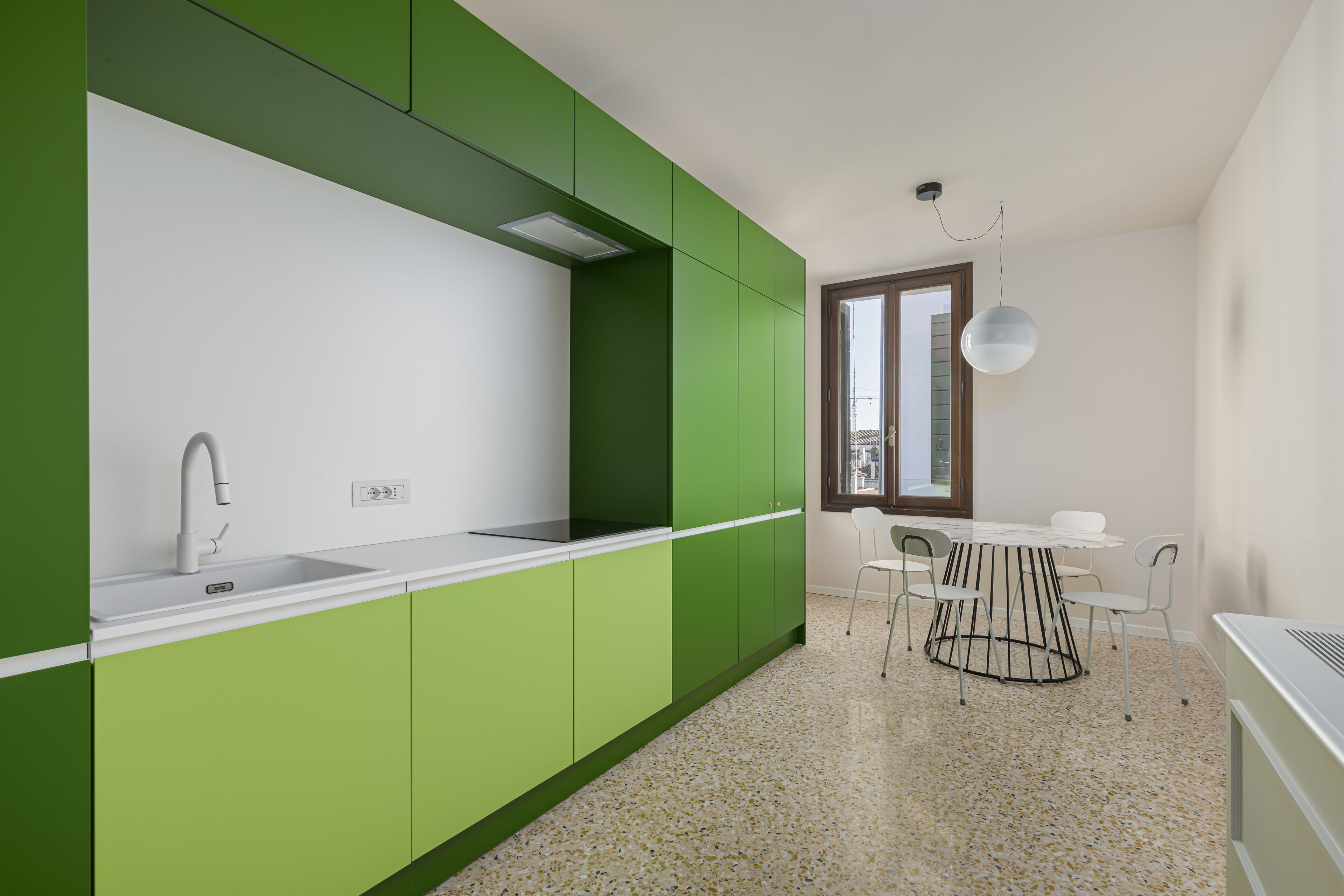 Une cuisine moderne avec des meubles verts, un plan de travail blanc et un évier. On y trouve un coin repas avec une table et des chaises, ainsi qu’une fenêtre donnant sur un beau paysage.